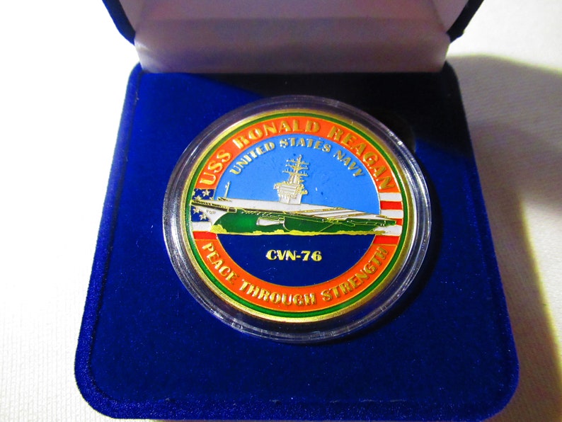Op de afbeelding: Een gouden munt met een blauw, groen en rood ontwerp. De munt toont een afbeelding van de vliegdekschip USS Ronald Reagan en de tekst "USS Ronald Reagan United States Navy CVN-76 Peace Through Strength".