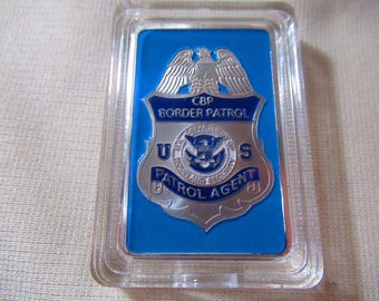 Könnte beinhalten: Ein blaues und silbernes Plastikabzeichen mit dem Text "CBP Border Patrol U.S. Patrol Agent" und einem Adler-Design.