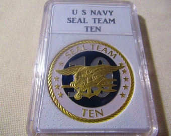 May include: A gold-colored coin with a black and white design. The coin features a Navy SEAL emblem with a trident and the words "SEAL TEAM TEN".