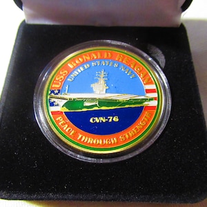 Op de afbeelding: Een gouden munt met een blauw, groen en oranje ontwerp. De munt toont een afbeelding van de USS Ronald Reagan vliegdekschip en de tekst "USS Ronald Reagan United States Navy CVN-76 Peace Through Strength".