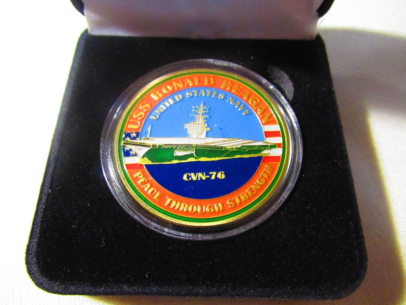 Op de afbeelding: Een gouden munt met een blauw, groen en oranje ontwerp. De munt toont een afbeelding van de USS Ronald Reagan vliegdekschip en de tekst "USS Ronald Reagan United States Navy CVN-76 Peace Through Strength".
