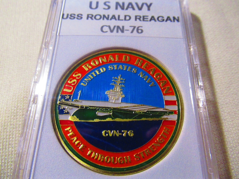 Op de afbeelding: Een gouden munt met een blauw, wit en rood ontwerp. De munt toont een afbeelding van een marinevliegdekschip met de tekst "USS RONALD REAGAN" en "CVN-76". De tekst "UNITED STATES NAVY" en "PEACE THROUGH STRENGTH" is ook zichtbaar op de munt.