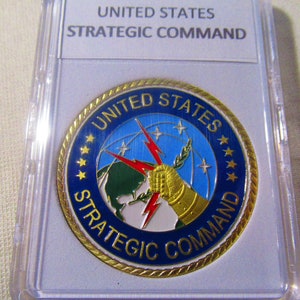 Puede incluir: Una moneda de color dorado con un diseño azul, blanco y verde. La moneda presenta las palabras "United States Strategic Command" y una imagen estilizada de un globo terráqueo con rayos y una mano sosteniendo una rama.