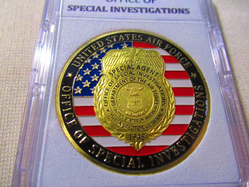 Może przedstawiać: Złoty medalion z czarnym obramowaniem i napisem "United States Air Force Office of Special Investigations" wok&oacute;ł krawędzi. W centrum medalionu znajduje się złota odznaka z napisem "Special Agent" i "Office of Special Investigations Department of the Air Force" wok&oacute;ł krawędzi. Odznaka zawiera r&oacute;wnież stylizowanego orła i napis "The Inspector General 1948".