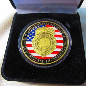 Może przedstawiać: Złoty medal z czerwono-biało-niebieskim wzorem amerykańskiej flagi. Medal zawiera napis "United States Air Force" i "Office of Special Investigations" wok&oacute;ł krawędzi. W centrum medalu znajduje się złota odznaka z napisem "Special Agent" i "Department of the Air Force".