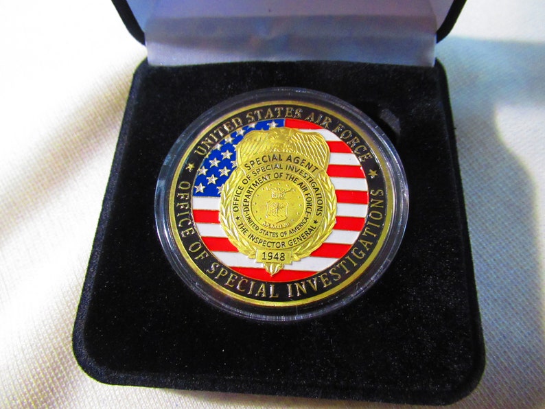 Może przedstawiać: Złoty medal z czerwono-biało-niebieskim wzorem amerykańskiej flagi. Medal zawiera napis "United States Air Force" i "Office of Special Investigations" wok&oacute;ł krawędzi. W centrum medalu znajduje się złota odznaka z napisem "Special Agent" i "Department of the Air Force".