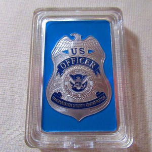 May include: A silver and blue US Transportation Security Administration officer badge in a clear plastic display case.