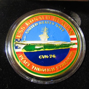 Op de afbeelding: Een gouden munt met een rood, wit en blauw Amerikaans vlagontwerp. De munt toont een afbeelding van de USS Ronald Reagan vliegdekschip met de tekst "USS Ronald Reagan United States Navy CVN-76 Peace Through Strength".