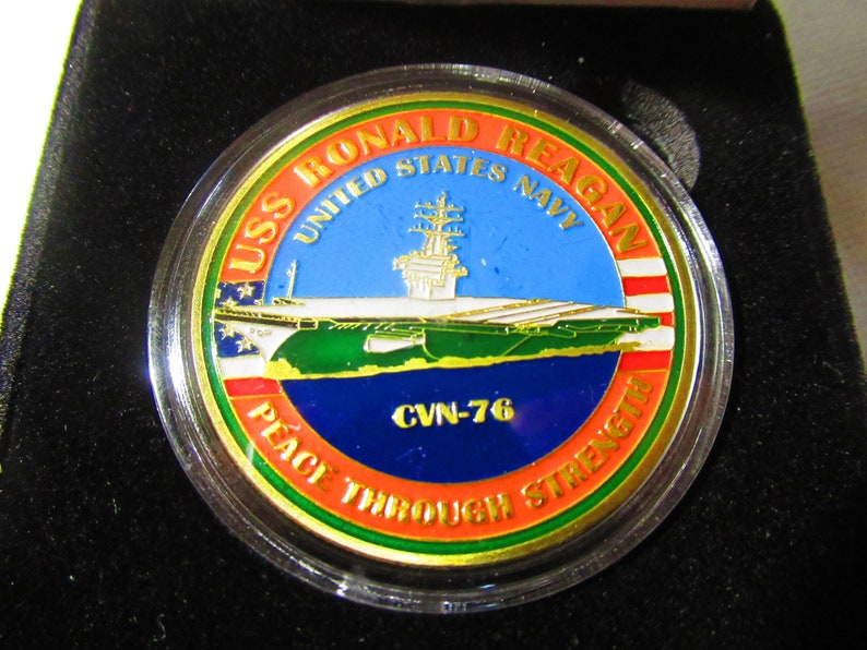 Op de afbeelding: Een gouden munt met een rood, wit en blauw Amerikaans vlagontwerp. De munt toont een afbeelding van de USS Ronald Reagan vliegdekschip met de tekst "USS Ronald Reagan United States Navy CVN-76 Peace Through Strength".