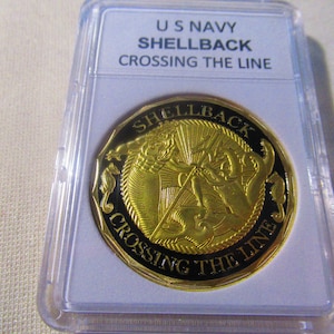 Può includere: Una moneta dorata con sfondo nero. La moneta presenta una raffigurazione di un marinaio che viene immerso nell'oceano da una creatura marina. Il testo "US NAVY SHELLBACK CROSSING THE LINE" è stampato sulla moneta.