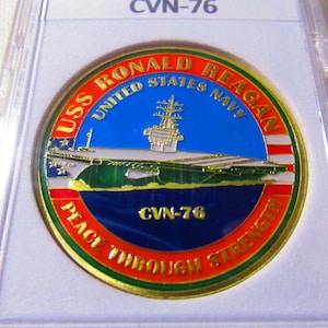 Op de afbeelding: Een gouden munt met een blauw, groen en rood ontwerp. De munt toont een afbeelding van de vliegdekschip USS Ronald Reagan en de tekst "USS Ronald Reagan United States Navy CVN-76 Peace Through Strength".
