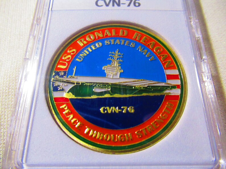 Op de afbeelding: Een gouden munt met een blauw, groen en rood ontwerp. De munt toont een afbeelding van de vliegdekschip USS Ronald Reagan en de tekst "USS Ronald Reagan United States Navy CVN-76 Peace Through Strength".