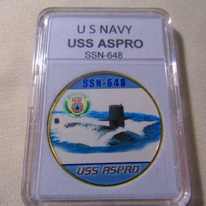 Puede incluir: Una moneda redonda con borde dorado con una imagen azul y blanca de un submarino que emerge en el océano. La moneda tiene el texto "USS ASPRO" y "SSN-648" en ella. La moneda está en una caja de plástico transparente.
