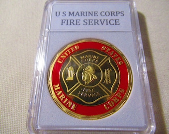 May include: A gold-colored US Marine Corps Fire Service coin encased in a clear plastic holder. The coin features a red and gold design with the words "US MARINE CORPS FIRE SERVICE" at the top.