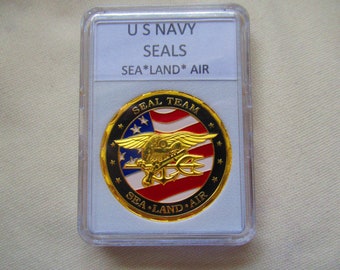 Pode incluir: Uma moeda dourada com uma borda preta e um design de bandeira americana vermelha, branca e azul. A moeda apresenta as palavras "SEAL TEAM" e "SEA LAND AIR" ao redor da borda. A moeda está em uma caixa de plástico transparente com um rótulo branco que diz "US NAVY SEALS SEA*LAND*AIR".