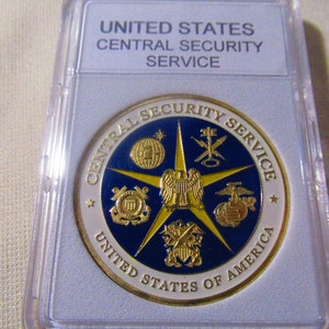 May include: A gold-colored coin with a blue and gold design. The coin features the words "Central Security Service" and "United States of America" around the edge. The center of the coin features a five-pointed star with the seals of the five branches of the United States Armed Forces.
