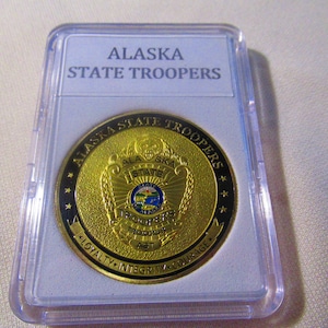 May include: A gold-colored coin with the words "Alaska State Troopers" around the edge. The center of the coin features a shield with the words "Alaska State" and a depiction of a mountain range and a star. The bottom of the coin reads "Loyalty, Integrity, Courage".
