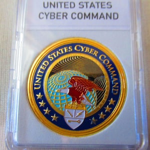 Puede incluir: Una moneda dorada con el texto "UNITED STATES CYBER COMMAND" en un borde azul y dorado. La moneda presenta un diseño de águila, globo terráqueo y escudo. La moneda está dentro de un soporte de plástico transparente.