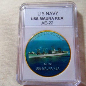 May include: A gold-colored coin with a blue and white image of the USS Mauna Kea, a US Navy ship. The coin has the text "United States Navy" and "AE-22 USS Mauna Kea" on it.