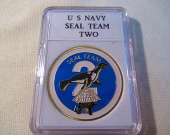 May include: A US Navy SEAL Team Two challenge coin in a plastic display case. The coin features a blue background with a white seal and the number 2. The seal is holding a parachute and a gun.
