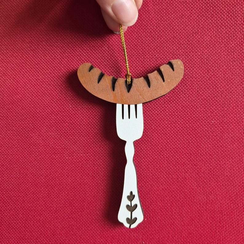 May include: A flat, wooden hanging ornament shaped like a white fork with a brown sausage resting on its tines. The sausage features dark brown grill marks, and the fork handle is decorated with a small, dark brown leaf-like pattern at the base.