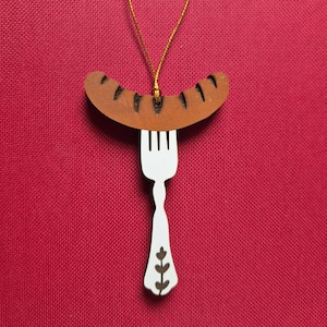 May include: A hanging ornament featuring a brown sausage with dark charred markings resting on a white fork. The fork handle has a decorative brown leaf pattern. A thin gold string is attached to the top of the sausage for hanging purposes.