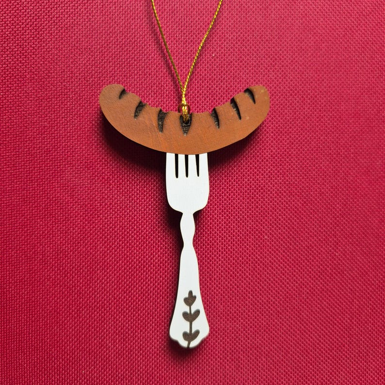 May include: A hanging ornament featuring a brown sausage with dark charred markings resting on a white fork. The fork handle has a decorative brown leaf pattern. A thin gold string is attached to the top of the sausage for hanging purposes.