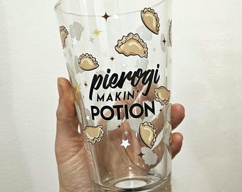 May include: A clear glass tumbler featuring a repeating pattern of beige pierogi illustrations, small white and yellow stars, and the black printed text "pierogi MAKIN' POTION" centred on the front surface of the drinking vessel.