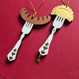 May include: Two hanging ornaments shaped like white forks. One fork holds a brown sausage, and the other holds a gold-colored pierogi. Both ornaments feature decorative cutouts on the handles, and the pierogi ornament includes a small gold star charm.