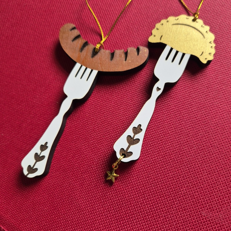 May include: Two hanging ornaments shaped like white forks. One fork holds a brown sausage, and the other holds a gold-colored pierogi. Both ornaments feature decorative cutouts on the handles, and the pierogi ornament includes a small gold star charm.