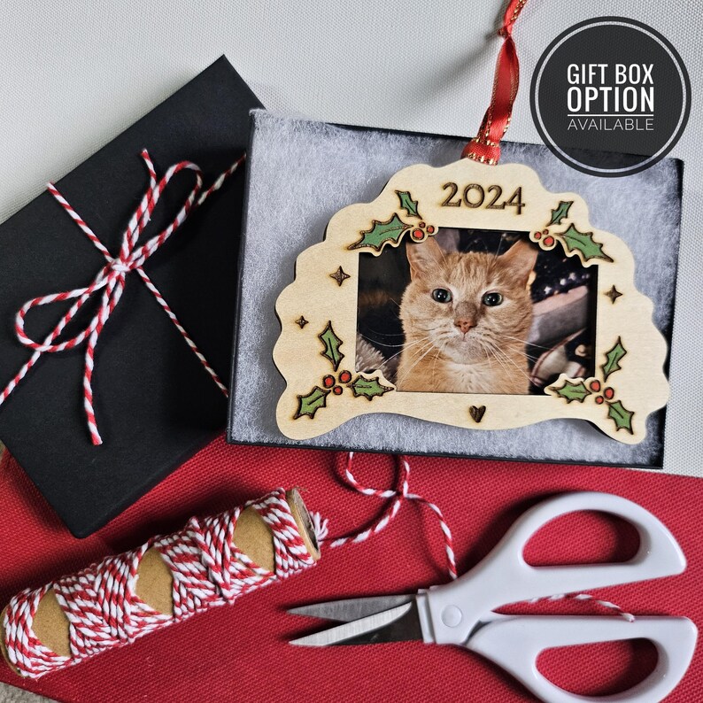 May include: A light-colored wooden picture frame ornament featuring a photo of an orange cat, engraved with the year 2024 and decorated with green holly leaves and red berries. The ornament includes a red ribbon and is presented in a black box.