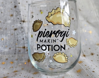 May include: A clear glass wine glass with a gold and black design. The glass features a graphic of pierogies and the text "pierogi makin' potion".