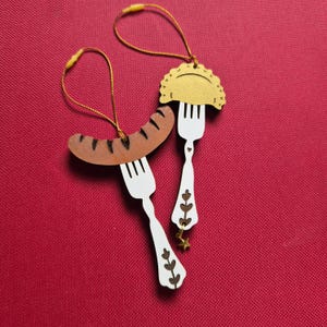 May include: Two hanging ornaments featuring white forks with brown floral cutouts. One fork holds a brown sausage with dark grill marks, and the other holds a golden pierogi. Both ornaments include gold hanging strings and a small gold star charm.