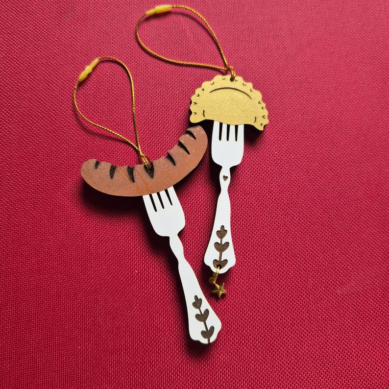 May include: Two hanging ornaments featuring white forks with brown floral cutouts. One fork holds a brown sausage with dark grill marks, and the other holds a golden pierogi. Both ornaments include gold hanging strings and a small gold star charm.