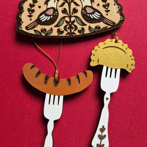 May include: A set of three wooden hanging ornaments featuring a semi-circular plaque with two birds and floral motifs, a brown sausage on a white fork, and a gold-colored dumpling on a white fork with a small star charm attached to the handle.
