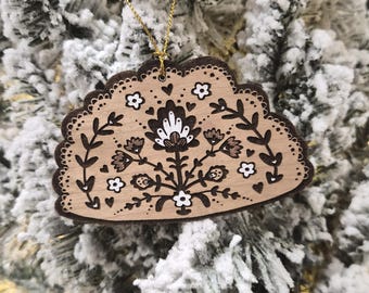 May include: A semi-circular wooden ornament featuring a dark brown laser-cut edge and a detailed floral design in black and white ink. The ornament includes a small hole at the top with a thin gold string attached for hanging on a tree branch.