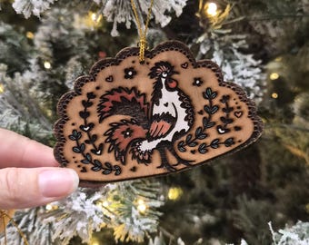 May include: A semi-circular wooden ornament featuring a pyrography illustration of a rooster surrounded by floral motifs, hearts, and small stars. The natural wood piece has a scalloped edge and is suspended by a thin gold string for hanging.