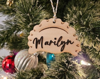 May include: A light-colored wooden ornament shaped like a pierogi, featuring the name Marilyn laser-cut in dark script lettering. The ornament has decorative etched lines along the curved edge and hangs from a white and gold twisted string.