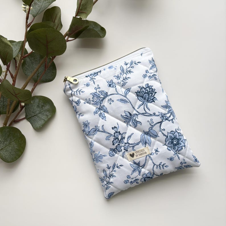 Może przedstawiać: Biała pikowana kosmetyczka z zamkiem błyskawicznym, z niebieskim wzorem kwiatowym i napisem "Stitched by Tashiana".