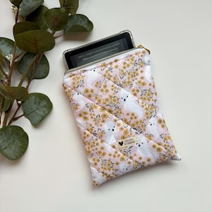 May include: A pink quilted sleeve with a floral and ghost pattern, holding an e-reader. The sleeve has a gold zip and a tag that reads "Stitched by Tabitha."