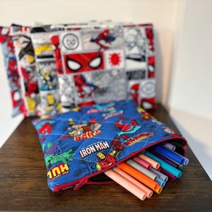 May include: A quilted fabric pouch featuring a blue background with Marvel comic book character graphics including Iron Man, Thor, and Hulk, with a red zip closure containing various coloured plastic markers or pens, and additional pouches in the background.