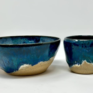 Peut inclure: Un ensemble de bol et de tasse en céramique. Le bol est large et peu profond, et la tasse est cylindrique. Les deux pièces ont un glaçage bleu foncé brillant qui s'estompe en bleu clair et beige près de la base.