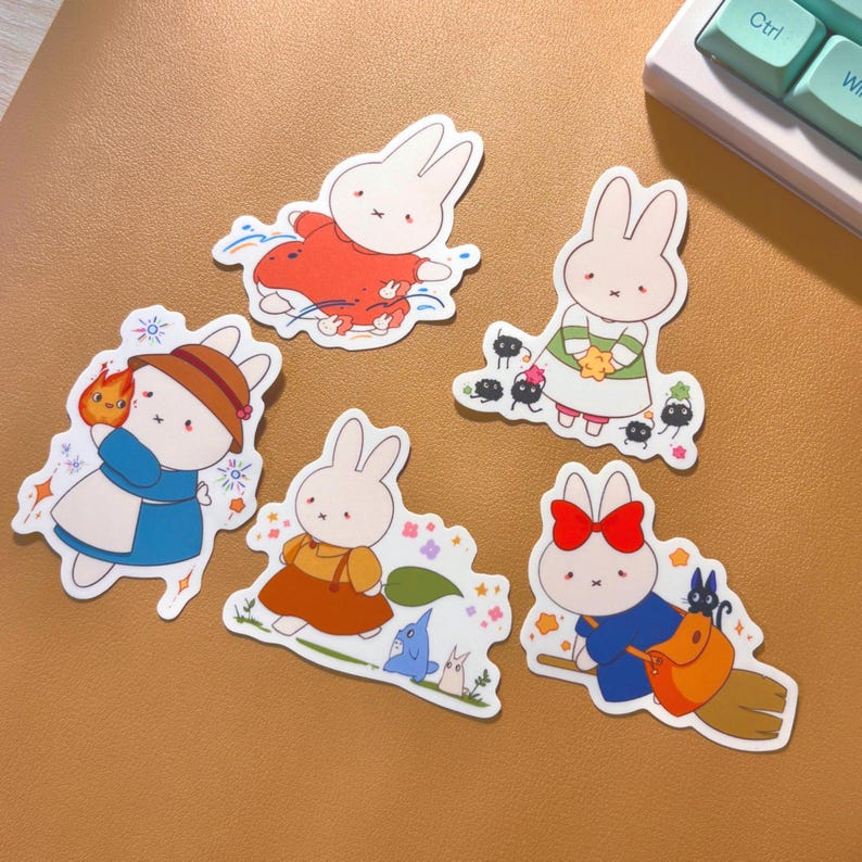 Op de afbeelding: Vijf kleurrijke stickers met cartoon-konijnen in verschillende poses. De stickers zijn gedrukt op wit papier en hebben een glanzende afwerking. De konijnen dragen verschillende outfits, waaronder een rode jurk, een blauwe jurk, een groene jurk, een bruine jurk en een blauw-witte jurk. De stickers zijn perfect om een vleugje whimsy toe te voegen aan je planner, laptop of waterfles.