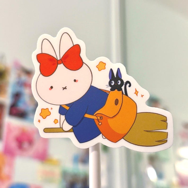 Op de afbeelding: Een witte sticker met een cartoonkonijn dat een blauwe en oranje outfit draagt met een rode strik, rijdend op een bezem met een zwarte kat op zijn rug. Het konijn heeft een roze neus en een gestikte mond.