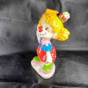 Pu&ograve; includere: Statuetta di clown in ceramica con capelli gialli, naso rosso e cappello da festa. Il clown indossa un abito rosa e blu con pois rossi e scarpe verdi. La statuetta poggia su una base bianca con bordi rosa.