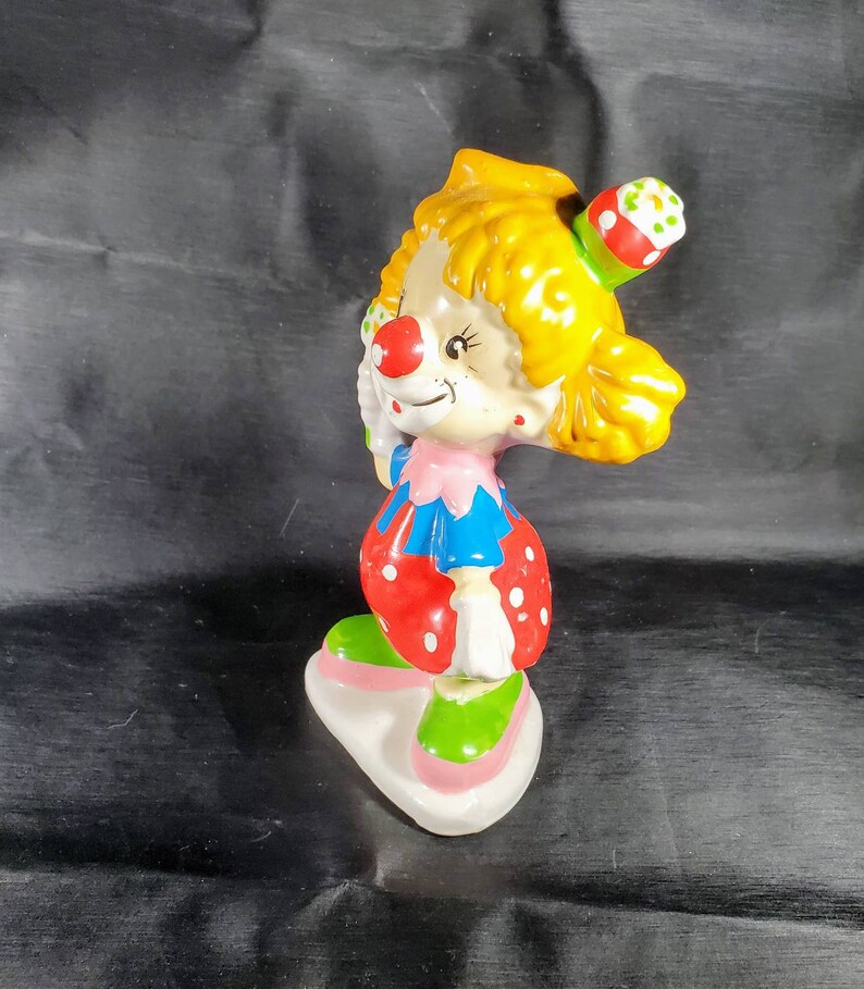 Pu&ograve; includere: Statuetta di clown in ceramica con capelli gialli, naso rosso e cappello da festa. Il clown indossa un abito rosa e blu con pois rossi e scarpe verdi. La statuetta poggia su una base bianca con bordi rosa.