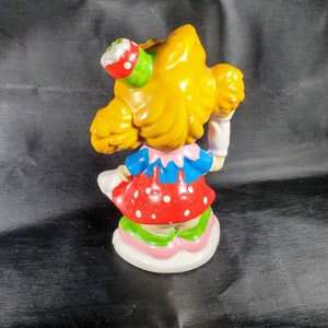 Pu&ograve; includere: Una statuina in ceramica di una ragazza con i capelli gialli, che indossa un top blu, un vestito rosso a pois e una gonna multicolore. La statuina tiene in mano un piccolo oggetto sulla testa ed &egrave; in piedi su una base bianca.