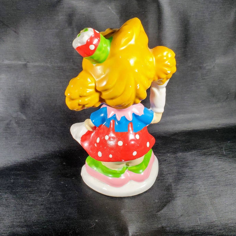 Pu&ograve; includere: Una statuina in ceramica di una ragazza con i capelli gialli, che indossa un top blu, un vestito rosso a pois e una gonna multicolore. La statuina tiene in mano un piccolo oggetto sulla testa ed &egrave; in piedi su una base bianca.