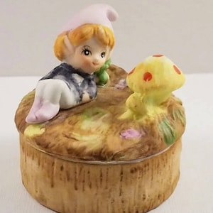 Puede incluir: Caja de baratijas de cerámica con un diseño caprichoso. La tapa está adornada con una figura de elfo sentado con un sombrero rosa y una seta. La caja tiene una textura marrón claro, similar a la madera.