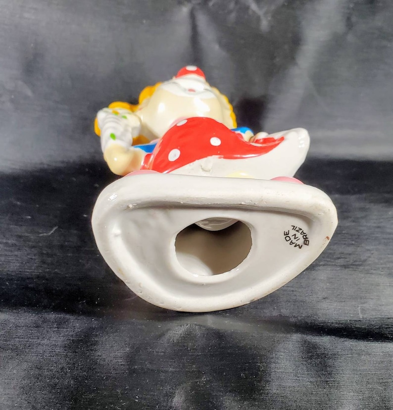 Pu&ograve; includere: Statuetta di clown in ceramica con cappello rosso a pois bianchi e base bianca. Il clown ha i capelli gialli, un abito blu e rosso e tiene in mano un oggetto colorato. La statuetta &egrave; contrassegnata "Made in Brazil".